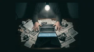 A person's hands splayed over a cluttered desk with fragmented news clippings and a laptop displaying garbled headlines, illuminated by a harsh overhead bulb.